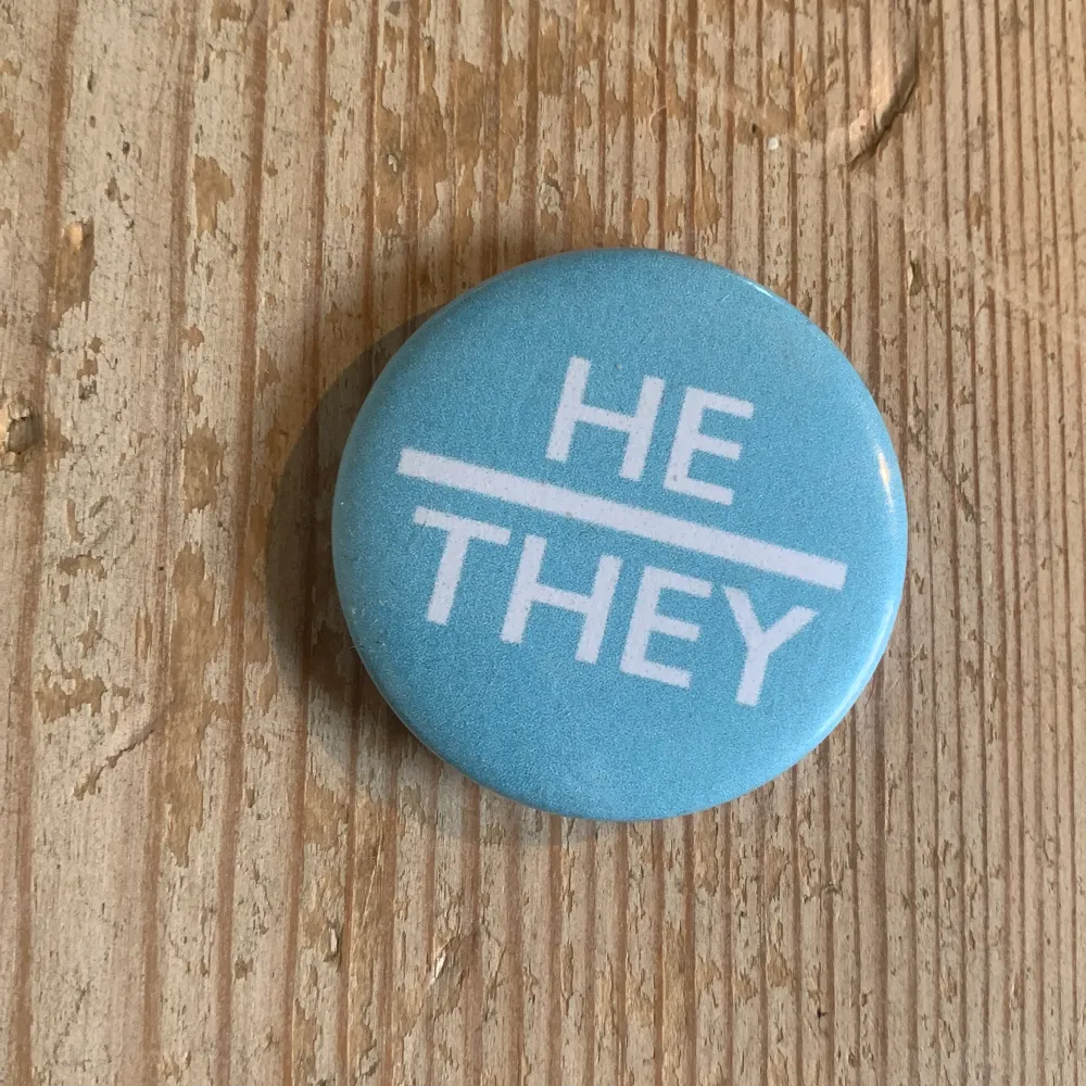 En rund knapp i ljusblå färg med vit text som säger 'HE/THEY'. Perfekt för att uttrycka pronomen på ett stilrent sätt. Knappen har en slät yta och en säkerhetsnål på baksidan för enkel fastsättning på kläder eller väskor.. Accessoarer.