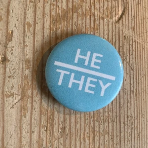 Blå knapp med texten 'HE/THEY' - En rund knapp i ljusblå färg med vit text som säger 'HE/THEY'. Perfekt för att uttrycka pronomen på ett stilrent sätt. Knappen har en slät yta och en säkerhetsnål på baksidan för enkel fastsättning på kläder eller väskor.