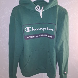 Grön hoodie från Champion - Säljer en snygg grön hoodie från Champion i storlek medium. Den har en stor logga på framsidan och en praktisk känguruficka. Perfekt för en avslappnad stil och passar bra till både höst och vår. Huvan har en vit dragsko för justering. Skön och bekväm att bära! Kommer aldrig till användning pga att jag inte passar i grönt💞