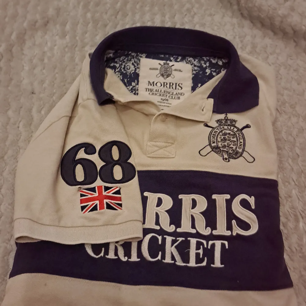 Säljer en snygg cricket t-shirt från Morris i beige och marinblått. Den har korta ärmar och ett klassiskt tryck med texten 'Morris Cricket' på framsidan. Perfekt för en avslappnad stil eller sportiga aktiviteter. Stor logga på bröstet och nummerdetalj på ärmen. Skön och stilren!💯💯. T-shirts.