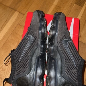 Svarta Nike Vapormax sneakers - Säljer ett par svarta Nike Vapormax sneakers i bra skick. De har en snygg och modern design med stickat material och en luftig känsla. Skorna har snörning och en genomskinlig sula som ger en cool look. Perfekta för både träning och vardag! Säljer en för billig slant då de kostar 2200 köpta med kvitto 🧾 och fårs med i paketet 