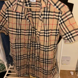 Rutig kortärmad skjorta från Burberry - Säljer en klassisk Burberry-skjorta i beige med det ikoniska rutmönstret. Skjortan är kortärmad och perfekt för både vardag och fest. Den är i bra skick och har alla knappar framtill. Den är i storlek xs men den passar mellan xs-small enligt mig. 