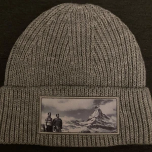Grå ribbad mössa med bergsmotiv - Snygg grå ribbad mössa med ett coolt bergsmotiv på framsidan. Perfekt för kalla dagar när du vill hålla dig varm och samtidigt se stilren ut. Tillverkad av mjukt material som känns skönt mot huden. Passar både till vardags och utomhusaktiviteter. Helt oanvänd.