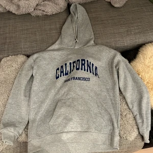 Grå hoodie med tryck 'California San Francisco' - Säljer en grå hoodie i storlek m med trycket 'California San Francisco' i mörkblått. Den är supermjuk och perfekt för kyliga dagar. Har en stor ficka fram och en bekväm huva. Perfekt för en avslappnad stil eller mysiga hemmakvällar. Använd hemma en gång 