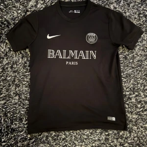 Svart t-shirt från Nike x Balmain - Säljer en snygg svart t-shirt från Nike i samarbete med Balmain. Den har en stilren design med Balmain Paris-logga och Nike-märke på framsidan. Perfekt för både vardag och träning. Materialet är Dri-FIT vilket gör den bekväm och andas bra. Passar perfekt till jeans eller träningsbyxor!