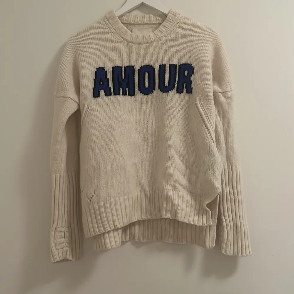 Säljer en mysig beige stickad tröja med blå och svart text 'AMOUR' på framsidan. Tröjan har långa ärmar och ribbade detaljer vid ärmslut och nederkant. Perfekt för kyliga dagar och ger en avslappnad stil. Endast lite slitage på framsidan, men fortfarande i bra skick.. Tröjor & Koftor.