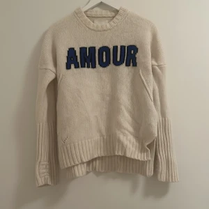 Amour stickad tröja - Säljer en mysig beige stickad tröja med blå och svart text 'AMOUR' på framsidan. Tröjan har långa ärmar och ribbade detaljer vid ärmslut och nederkant. Perfekt för kyliga dagar och ger en avslappnad stil. Endast lite slitage på framsidan, men fortfarande i bra skick.