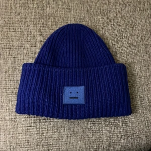 Blå stickad mössa från Acne Studios - Säljer en snygg blå stickad mössa från Acne Studios. Den har en ribbad design och ett coolt ansiktsmärke framtill. Perfekt för att hålla sig varm och stilren under höst och vinter. Passar de flesta tack vare one size-storleken. 🧢💙