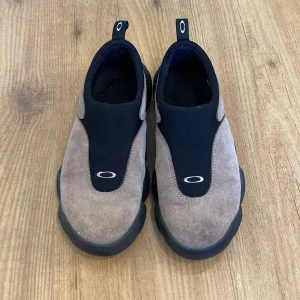 Bruna och svarta slip-on skor från Oakley - Säljer ett par snygga slip-on skor från Oakley i brunt och svart. Skorna har en stilren design med en bekväm passform och är perfekta för vardagsbruk. De är i mycket bra skick och har en robust sula för extra komfort. Perfekta för höstpromenader eller en avslappnad dag ute.
