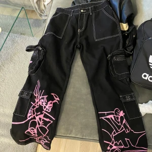 Emo Svarta cargobyxor med rosa tryck - Säljer ett par svarta cargobyxor med coolt rosa tryck. Byxorna har flera fickor med dragkedjor och en loose passform. Perfekta för en avslappnad stil med en edgy touch. Passar bra till både vardag och fest!