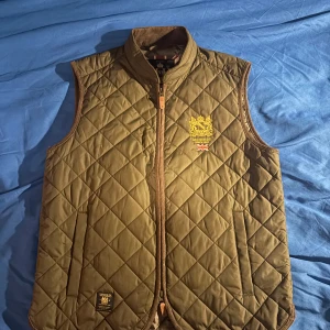 Olivgrön quiltad väst från Morris - Säljer en olivgrön quiltad väst från Morris i storlek medium. Västen har en snygg broderad logga på bröstet och en dragkedja framtill. Perfekt för höst och vår, med en klassisk och stilren design. Passar både till vardags och lite finare tillfällen. Använd ett fåtal gånger pågrund av att jag växte ur den. Original pris 1999kr, pris kan diskuteras!