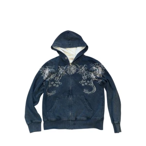 Ed Hardy Hoodie - Ed Hardy hoodie med dragkedja och drakmotiv på framsidan. Baksidan har en stor dödskalle med text och detaljerade mönster. Perfekt för en avslappnad stil med en edgy touch. Passar bra till höst och vår.