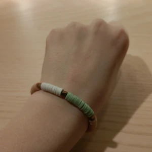 Färgglatt armband med pärlor - Säljer ett snyggt armband med platta pärlor i grönt, vitt och beige. Armbandet har små metalliska detaljer mellan pärlorna som ger det en extra touch. Perfekt för att liva upp vilken outfit som helst! Passar både till vardags och festliga tillfällen.🥂🐆🌸