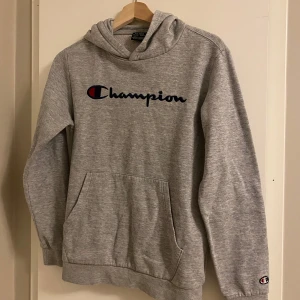 Grå hoodie från Champion  - Säljer en grå hoodie från Champion i storlek XL (barn). Den har en klassisk design med Champion-loggan broderad på bröstet och en liten logga på ärmen. Perfekt för höst och vinter, med en mysig känsla och känguruficka framtill! OBS den är i storlek XL för BARN