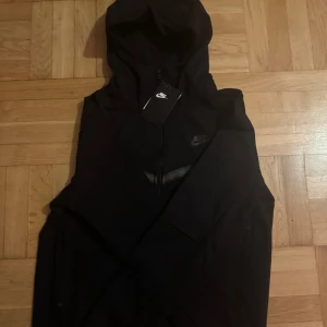 Svart träningsset från Nike - Säljer ett svart träningsset från Nike i mycket bra skick. Setet består av en långärmad hoodie med dragkedja och matchande byxor. Perfekt för träning eller en avslappnad dag. Materialet är mjukt och bekvämt, och det finns praktiska fickor med dragkedjor.