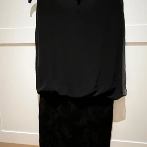 Svart klänning med spetsdetaljer - Elegant svart klänning med vackra spetsdetaljer på nederdelen. Den har en lös passform och är ärmlös, vilket gör den perfekt för festliga tillfällen. Materialet är lätt och luftigt, vilket ger en fin rörelse. Perfekt för en kväll ute eller en speciell tillställning.