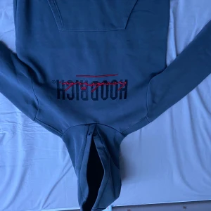 Grå hoodie från Hoodrich - Säljer en snygg grå hoodie från Hoodrich med coola röda detaljer och tryck både fram och bak. Den har en klassisk känguruficka och en bekväm passform. Perfekt för en avslappnad stil eller streetwear-look. Passar bra till både jeans och joggers! Obs!! Den har sliten del på luvan (Syns på bilden) annars är den fläckfri 