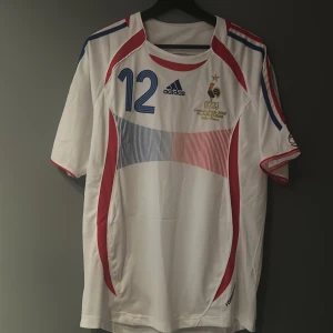 Henry tröja från Vm 2006 helt ny med tagg - Säljer en vit Adidas fotbollströja med nummer 12 och namntryck på baksidan. Tröjan har korta ärmar och detaljer i blått och rött. Den har ett broderat emblem på bröstet och är i mycket bra skick. Perfekt för samlare eller fans! Storlek L är en Replika och är väldigt bra kvalite helt ny med tagg