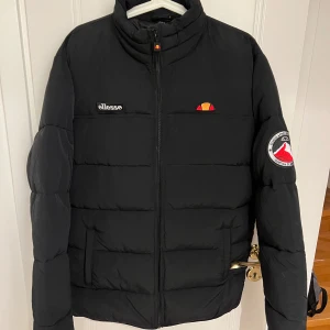 Svart pufferjacka från Ellesse - Säljer en svart pufferjacka från Ellesse i mycket bra skick. Jackan har en dragkedja framtill och är perfekt för kyliga dagar. Den har långa ärmar och en stilren design med Ellesse-loggan på bröstet och ärmen. Perfekt för både vardag och utomhusaktiviteter!
