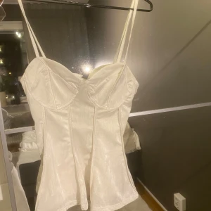 Vit korsett från H&M - Snygg vit korsett från H&M i storlek S. Den har smala axelband och en smickrande passform med en lätt glansig yta. Perfekt för festliga tillfällen eller som ett stilrent basplagg. Baktill finns en elastisk del för extra komfort.