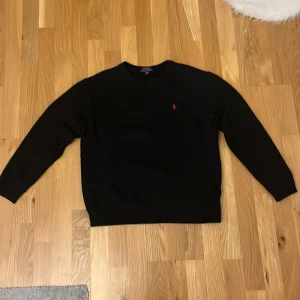 Svart tröja från Polo Ralph Lauren - Säljer en stilren svart tröja från Polo Ralph Lauren i storlek S. Den har en klassisk design med långärmade ärmar och en liten röd logga på bröstet. Perfekt för både vardag och festliga tillfällen. Tröjan är i mycket bra skick och redo för en ny ägare!