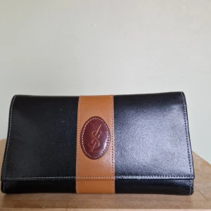 Svart och brun plånbok från Yves Saint Laurent (Äkthetsintyg från Tradera) - Vintage - Säljer en elegant svart och brun plånbok från Yves Saint Laurent. Den är tillverkad i skinn och har en stilren design med en brun rem och logotypdetalj. Insidan har en praktisk metallram för myntfack. Perfekt för att hålla ordning på dina kort och kontanter. Jag har haft den på Tradera och de har certifierat den som äkta. Se min andra annons på tillhörande väska.