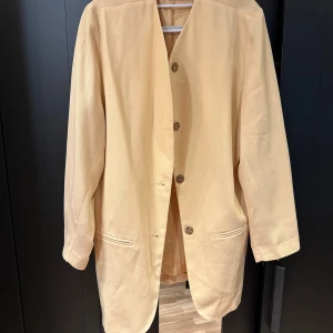 Beige kavaj - En stilren beige/guk kavaj i nyskick från Danmark i storlek 40! Den har en klassisk design med knappar framtill och långa ärmar. 