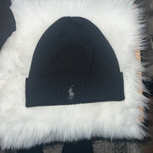 Svart mössa från Polo Ralph Lauren - Snygg svart mössa från Polo Ralph Lauren med deras ikoniska broderade logga framtill. Den är stickad och har en ribbad kant som ger en skön passform.! Unsex, onesize