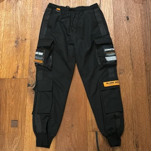 Svarta cargopants från Black Squad - Säljer ett par svarta cargopants från Black Squad i storlek XS. Byxorna har flera praktiska fickor med coola detaljer och text. De är perfekta för en avslappnad streetwear-look och har en bekväm passform med elastisk midja och snörning. Perfekta för höst och vår!