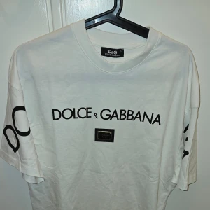 Vit t-shirt från Dolce & Gabbana Stl. L - Säljer en helt ny snygg vit t-shirt från Dolce & Gabbana med deras ikoniska logga tryckt i svart på framsidan i storlek L. T-shirten har en normal passform och är kortärmad, perfekt för en stilren och avslappnad look. Den är i nytt skick och passar både till vardags och fest. 