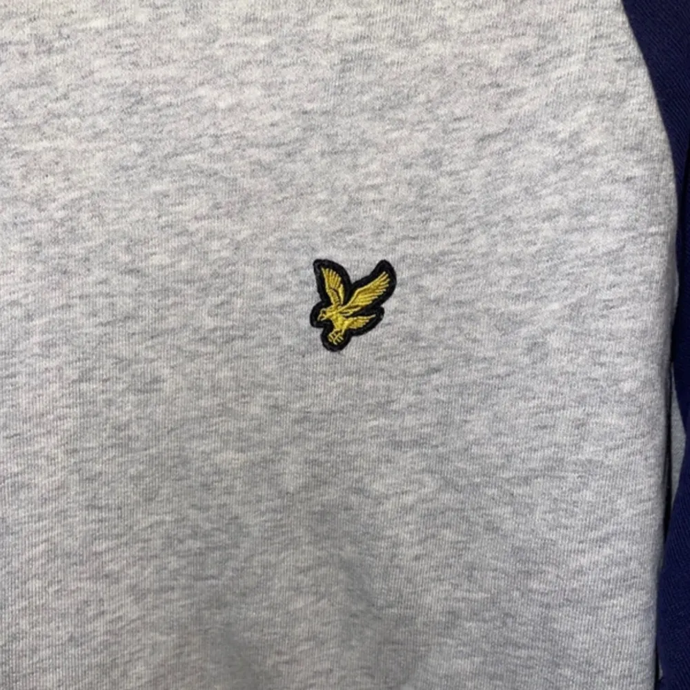 Säljer en snygg grå och marinblå tröja från Lyle & Scott. Den har ett broderat gult emblem på bröstet och långa ärmar. Perfekt för en avslappnad stil under höst och vår. Tröjan är i bra skick och redo för nya äventyr!. Tröjor & Koftor.