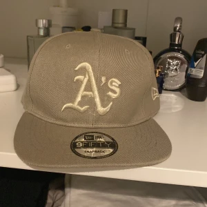 Beige keps från New Era - Säljer en beige snapback-keps från New Era med 'A's' broderat framtill. Kepsen har en justerbar rem baktill och MLB-logga. Perfekt för sommardagar eller som en stilren accessoar.