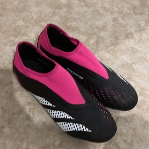 Adidas Predator fotbollsskor - Säljer ett par Adidas Predator fotbollsskor i svart och rosa. De har en strömlinjeformad design utan snörning och är perfekta för att dominera på planen. Skorna har en robust sula med rosa dobbar för optimalt grepp. Använda men i bra skick.