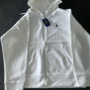 Vit hoodie från Polo Ralph Lauren - Säljer en stilren vit hoodie från Polo Ralph Lauren i storlek L. Den har en dragkedja framtill och en klassisk broderad logga på bröstet. Perfekt för en avslappnad och snygg look. Passar både till vardags och chillkvällar. 🏷️