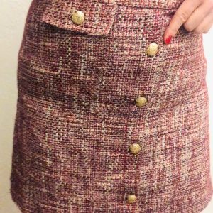 Rutig kjol med knappar - Snygg rutig kjol i en mix av röd, beige och brun toner. Kjolen har en rad med dekorativa guldknappar framtill och en liten fickdetalj upptill. Perfekt för höst och vinter, och kan stylas både upp och ner beroende på tillfälle. Passar bra till en stickad tröja eller en enkel blus.