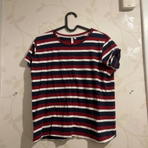 Randig t-shirt i bomull - Säljer en randig t-shirt i bomull med ränder i rött, vitt och blått. Den har en normal passform och korta ärmar, perfekt för sommardagar. T-shirten är i bra skick och redo för nya äventyr!