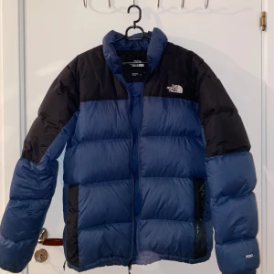 Blå och svart dunjacka från The North Face - Säljer en blå och svart dunjacka från The North Face. Jackan har en dragkedja framtill och är långärmad med ett klassiskt pufferdesign. Perfekt för kalla dagar.
