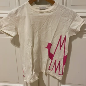 Vit t-shirt från Moncler med rosa tryck - Snygg vit t-shirt från Moncler med ett stort rosa tryck på framsidan och baksidan. T-shirten har korta ärmar och ett Moncler-märke på ärmen. Perfekt för en avslappnad stil. Barnstorlek 164/14y, skulle säga att det är en XS/S.