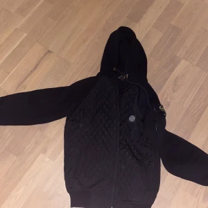 Svart cardigan från Stone Island - Snygg svart quiltad jacka med huva och dragkedja. Perfekt för kyligare dagar. Jackan har långa ärmar och en stilren design.