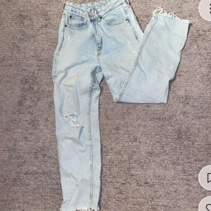 Ljusa jeans med slitningar - Säljer ett par ljusa jeans med slitningar och fransiga kanter. De har en klassisk femficksdesign och knappgylf. Perfekta för en avslappnad stil.