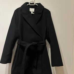 Svart kappa från H&M - Elegant svart kappa från H&M med knytskärp i midjan. Perfekt för kyligare dagar med sin stilrena design och långa ärmar. En klassisk och tidlös modell som passar till många tillfällen. Aldrig användes 