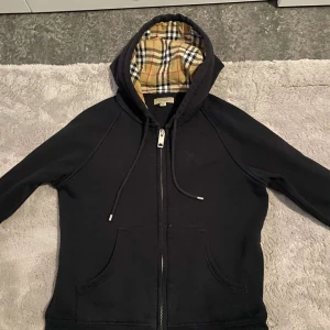 Svart huvtröja från Burberry - Säljer en stilren svart huvtröja från Burberry med klassiskt rutigt foder i huvan. Tröjan har dragkedja framtill. Tröjan är xs och växte ur den snabbt. Bra skipp, knappt använd