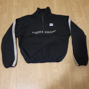 Svart tröja från Eivy - Säljer en svart tröja från Eivy med texten 'UNBORED ONBOARD' på bröstet och längs ärmarna. Tröjan har en halv dragkedja och är perfekt för en avslappnad stil.