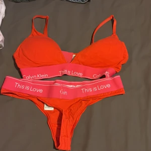 Röd underklädesset från Calvin Klein - Snyggt rött underklädesset från Calvin Klein med rosa resårband där det står 'This is Love'. Setet består av en bh och matchande trosor. Perfekt för en färgglad och bekväm stil.