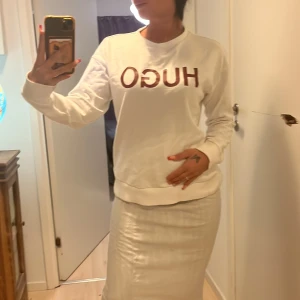 Sweatshirt HUGO BOSS - Säljer en stilren vit sweatshirt från Hugo med röd text på framsidan. Tröjan har långa ärmar och en röd rand med text på ryggen. Perfekt för en avslappnad stil.