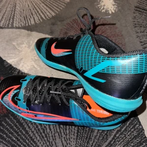Svarta och turkosa inomhus/fotbollskor från Nike - Snygga fotbollsskor/inomhusskor från Nike i svart och turkos med orange detaljer. Skorna har snörning och en sportig design som passar perfekt för fotbollsträning/inomhusträning. Dessa är i storlek 44 helt oanvända.