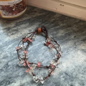 Armband med rosa och klara stenar - Unikt armband med rosa och klara stenar på en brun tråd. Armbandet har en naturlig och bohemisk stil med en enkel knäppning. Perfekt för att ge en färgklick till din outfit.