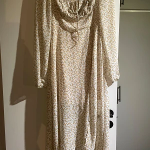 Blommig långklänning från ADOORE - Charmig långklänning från ADOORE med ett sött blommönster i gult och grönt på en vit bakgrund. Klänningen har långa ärmar och en knytning vid bysten för en romantisk touch. Perfekt för somriga dagar! Kommer aldrig till användning här tyvärr! 🌼