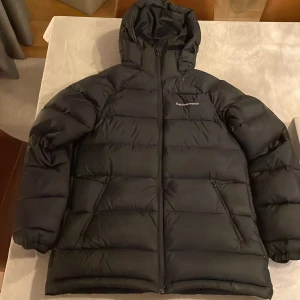 Svart dunjacka från Peak Performance - Säljer en svart dunjacka från Peak Performance med huva och dragkedja. Jackan är perfekt för kalla dagar och har en stilren design med logotyp på bröstet. Den är långärmad och har en pufferstil som ger extra värme.