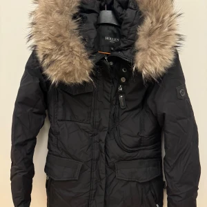 Svart parkajacka från Hollies - Snygg svart parkajacka från Hollies med en stor, fluffig pälsdetalj på huvan. Jackan har praktiska fickor och stängs med dragkedja och knappar. Perfekt för kyliga dagar.